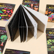 Open scratch art book with colorful designs on a beige surface, surrounded by closed books.